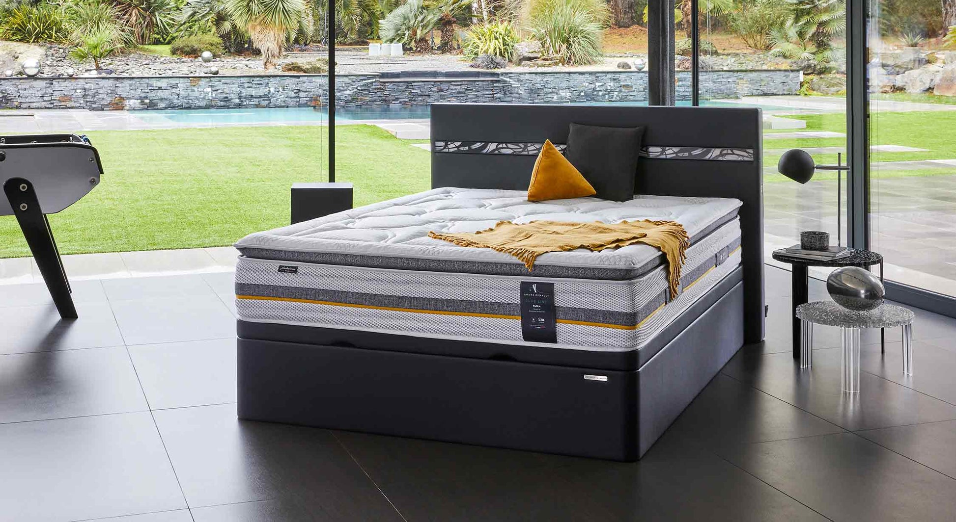 Lit coffre moderne avec matelas épais, décoré de coussins et plaid, dans une pièce lumineuse avec vue sur jardin et piscine