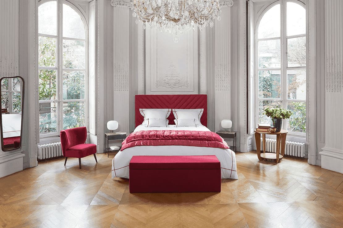 Chambre élégante avec lit et fauteuil rouges, parquet en bois, grandes fenêtres et lustre, créant une ambiance raffinée
