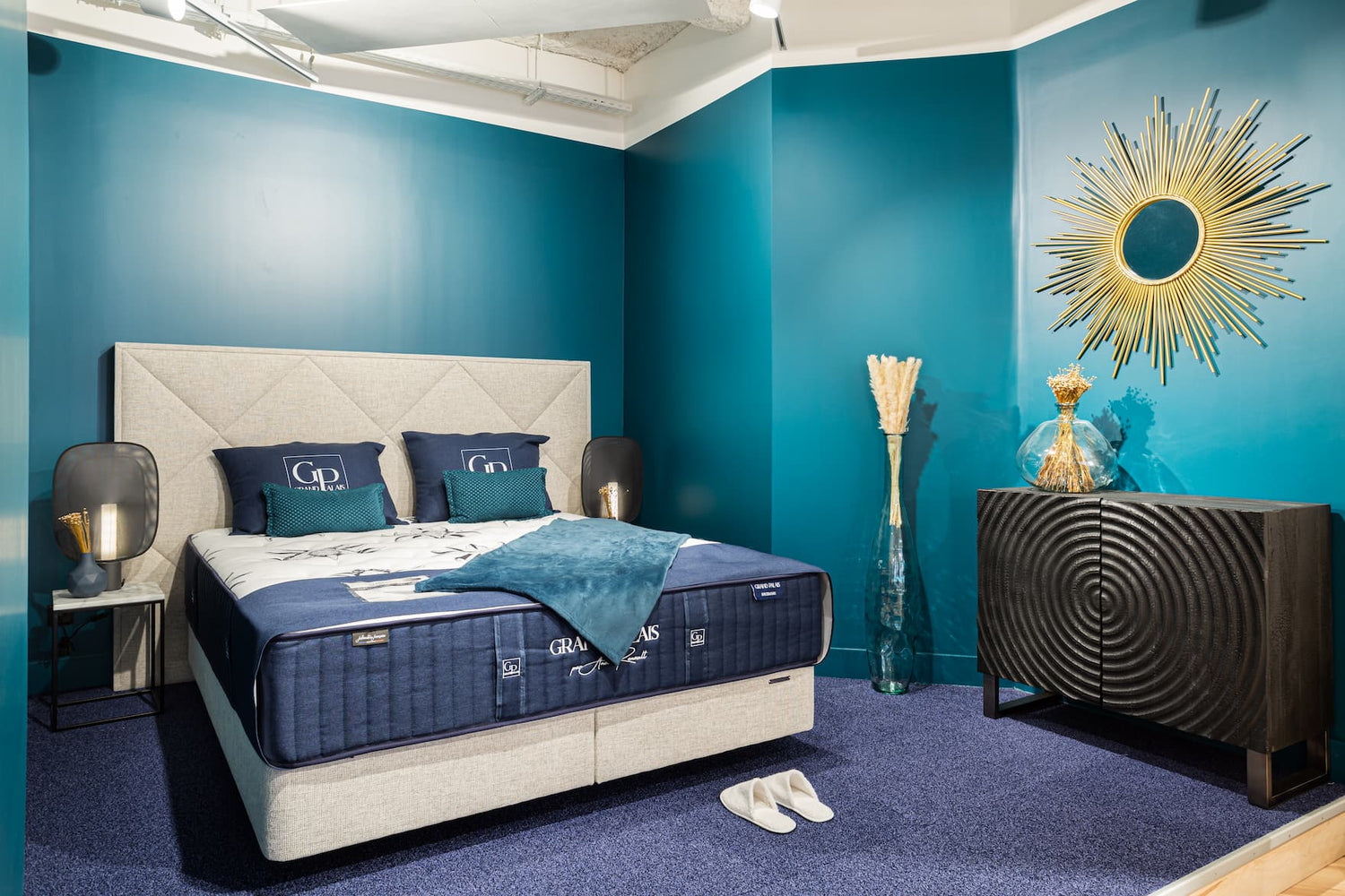 Chambre moderne avec lit double, coussins bleus, mur turquoise, miroir soleil doré et meuble noir texturé