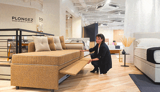 Lit gigogne beige avec tiroir dépliable, présenté par une femme dans un showroom moderne