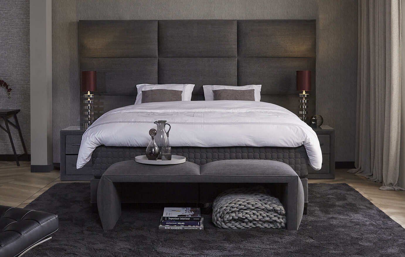 Chambre moderne avec grand lit, tête de lit capitonnée, banc gris, tapis foncé et décoration élégante