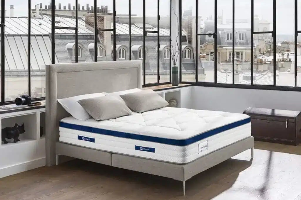 Matelas Simmons Gentleman dans une chambre moderne avec vue sur les toits parisiens, lit gris et grandes fenêtres