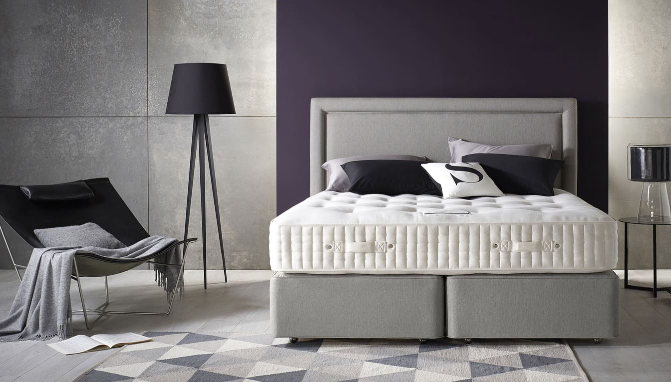 Chambre moderne avec lit rembourré, coussins noirs et blancs, lampe sur pied, fauteuil avec plaid gris, et mur violet