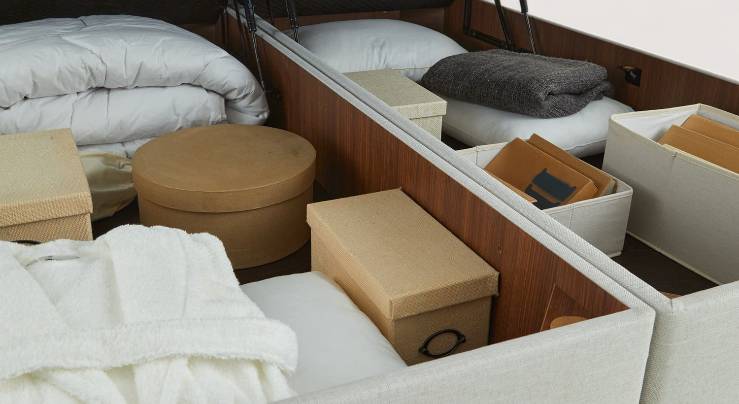 Espace de rangement sous un lit coffre en bois, contenant des boîtes de rangement, des couvertures blanches et des coussins