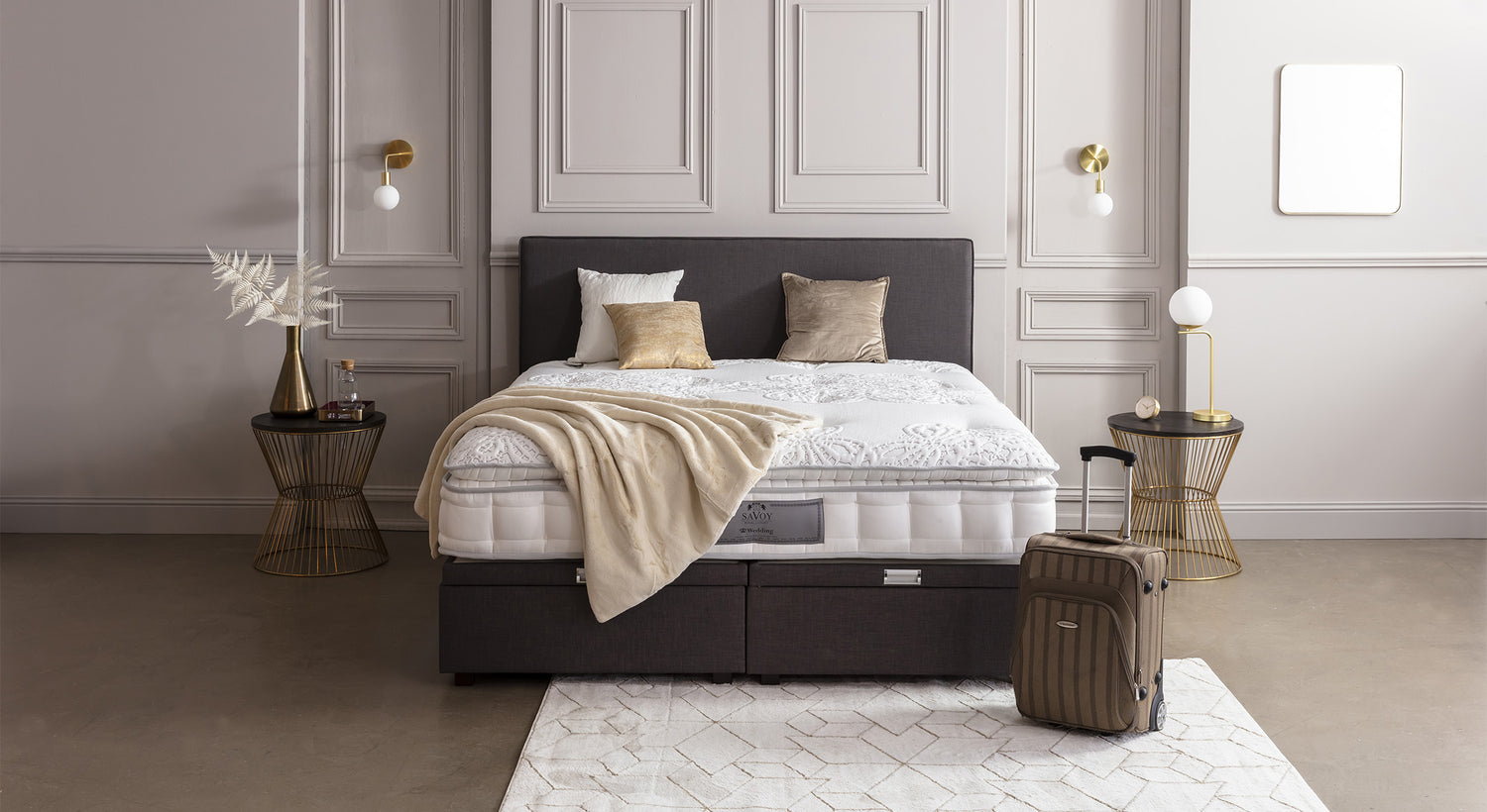 Lit coffre en tissu gris avec matelas, décoré de coussins et plaid beige, entouré de tables d'appoint dorées et valise à roulettes