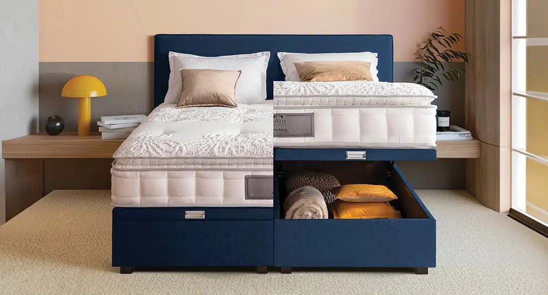 Lit coffre bleu avec matelas, tiroir ouvert contenant des couvertures et oreillers, dans une chambre moderne avec lampe jaune et plante