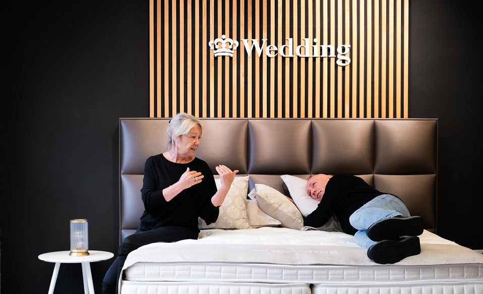 Deux personnes sur un lit, l'une assise et parlant, l'autre allongée. Panneau "Wedding" en arrière-plan. Contexte détente ou showroom