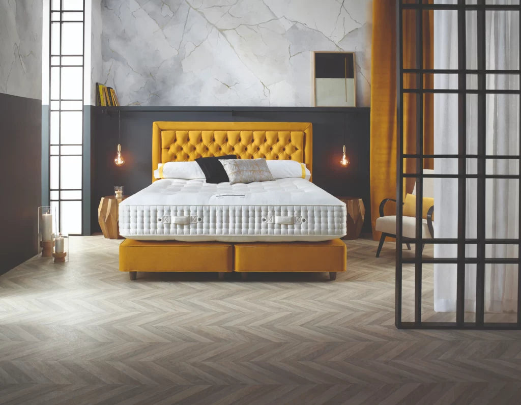 Chambre élégante avec lit à tête de lit jaune, éclairage suspendu et parquet en chevrons, créant une ambiance chaleureuse et moderne