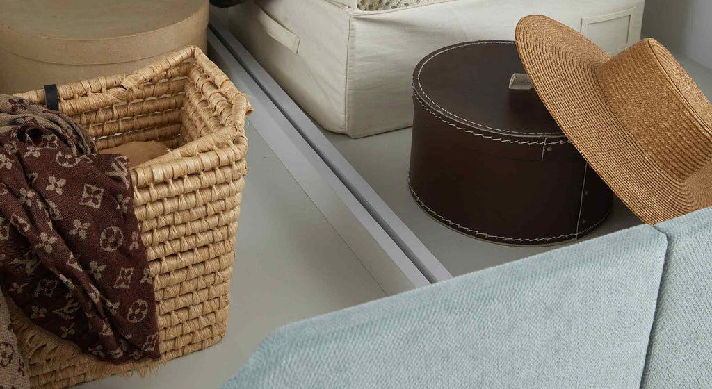 Panier en osier avec écharpe, boîte ronde en cuir, chapeau de paille et boîte en tissu sur un lit coffre