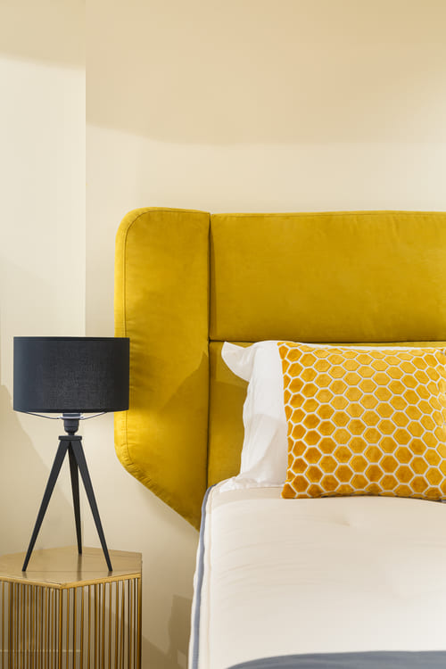 Lit avec tête de lit jaune, coussin à motifs hexagonaux, lampe noire sur table en bois. Ambiance moderne et chaleureuse