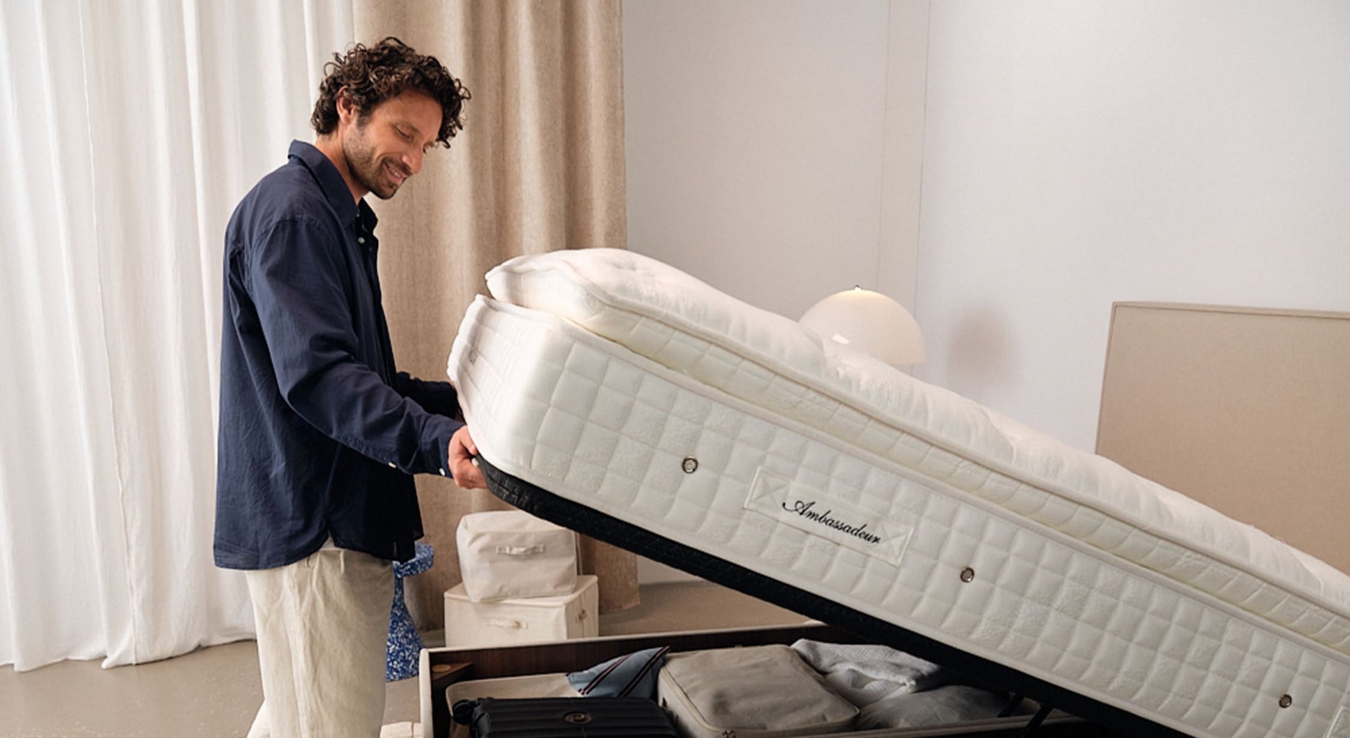 Homme soulevant le matelas d'un Lit coffre NINA Queens Beige pour accéder au rangement spacieux intégré, dans une chambre lumineuse et épurée.